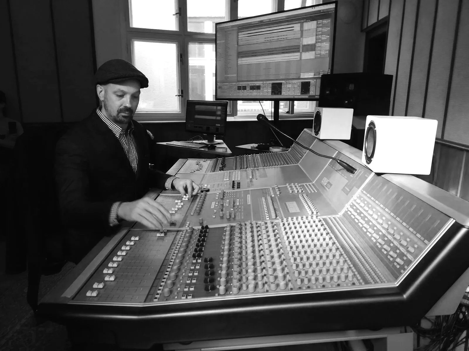 Black and white photo of a man in a hat working in a professional recording studio with large mixing console, multiple monitors, and studio speakers.