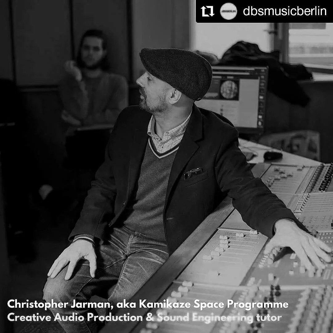 A black and white photo of Christopher Jarman, aka Kamikaze Space Programme, sitting at a sound mixing console in a recording studio. He is wearing a beret, a blazer, and has a beard. In the background, an individual is standing near a computer monitor and a window.
