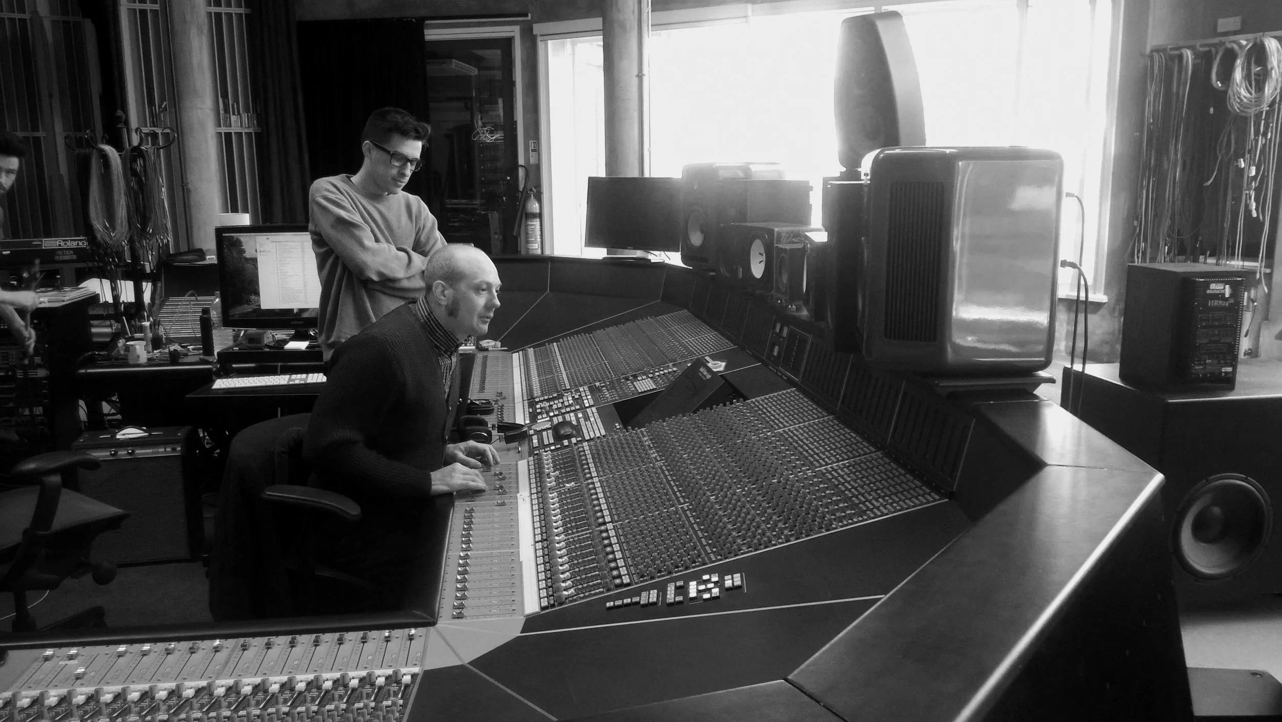 Two men working in a music recording studio, one seated at a large mixing console and the other standing behind him, with computer monitors, audio equipment, and large speakers in the background.