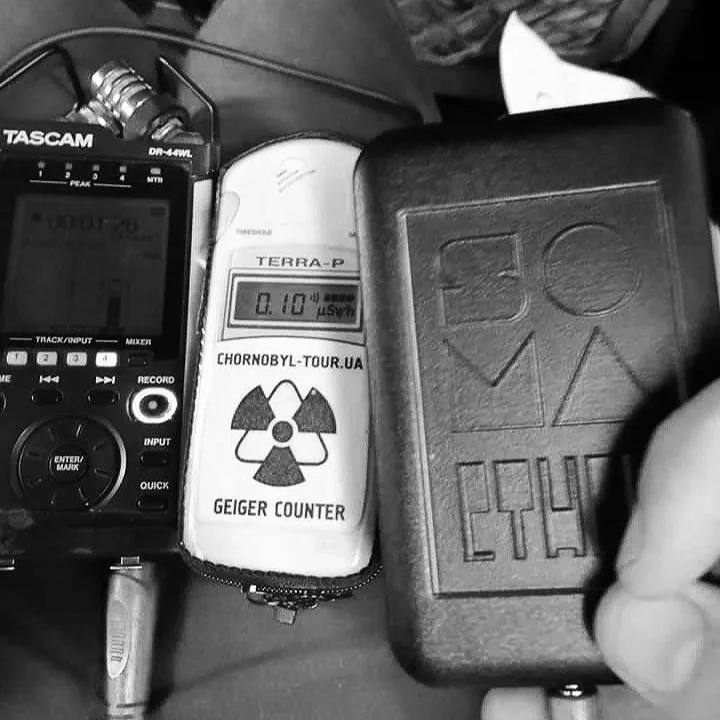 A radiation Geiger counter device with a display showing 0.10 microsieverts per hour, surrounded by a portable audio recorder, a notebook, and a zippered case, indicating a mobile monitoring setup.