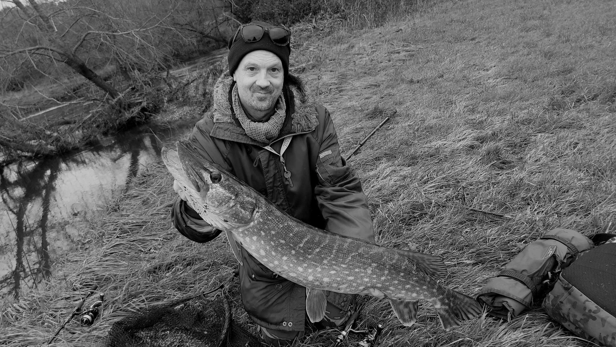 Man holding a large fish, standing outdoors near a small stream, with fishing gear and trees in the background, in black and white.