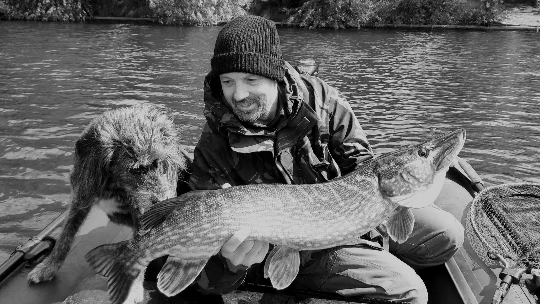A man in a camo jacket and beanie holding a large fish and a dog on a boat in a river