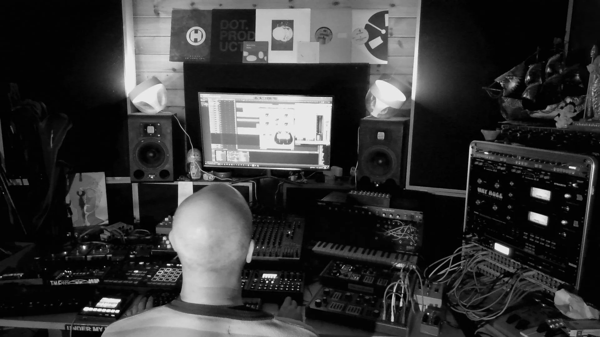 A person working in a music studio with electronic equipment, speakers, and a computer monitor displaying sound editing software, with posters on the wooden wall behind.