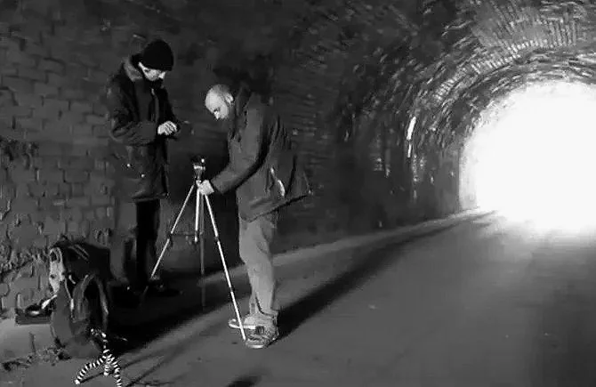 Two people in a tunnel, one adjusting a camera on a tripod, with a backpack and a dog nearby.