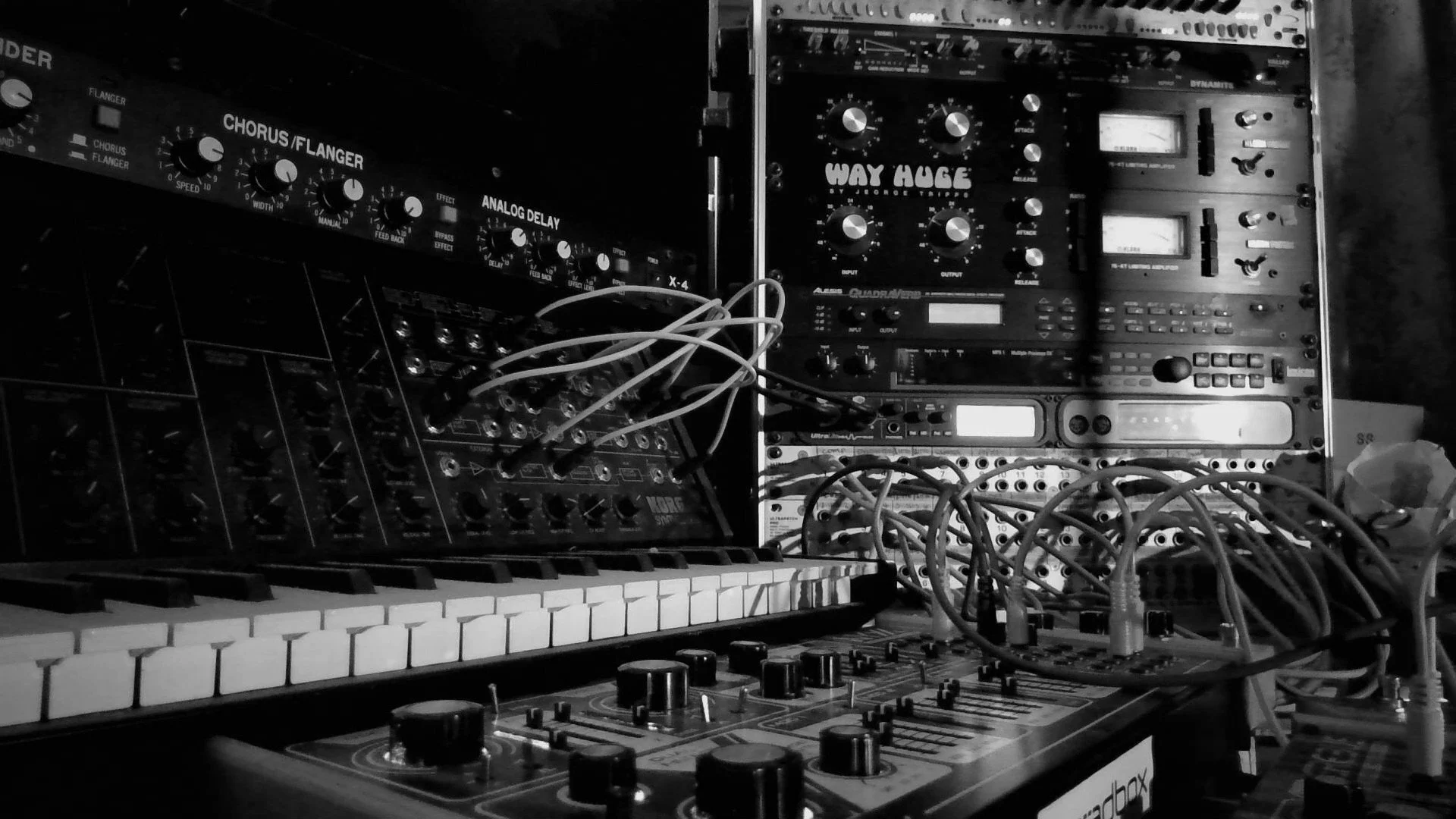 Black and white photograph of audio equipment, including a keyboard, mixers, audio processors, and various cables connected to the devices.