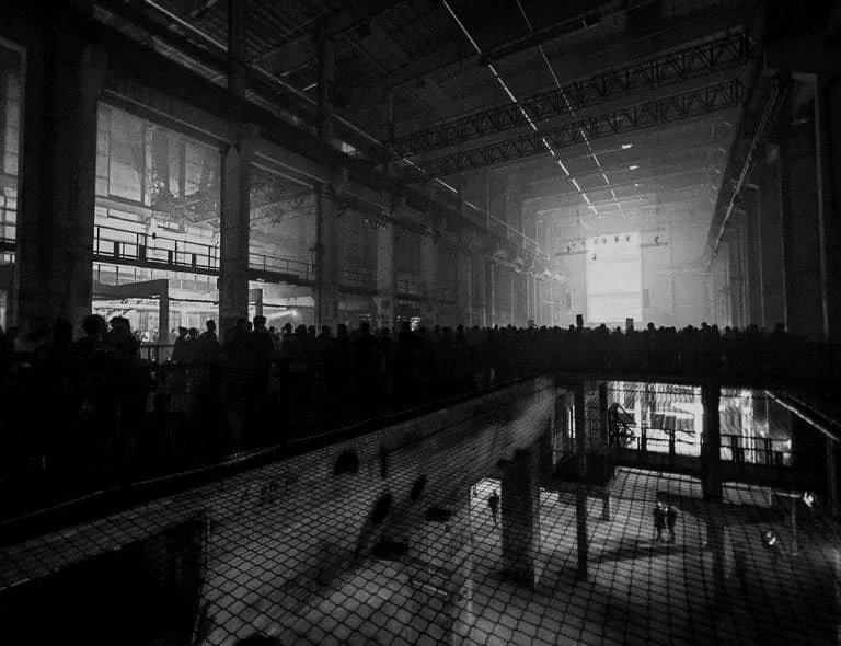 Inside a large, industrial-style building with high ceilings, crowds of people walking and standing, and a swimming pool with a mesh cover in the foreground.