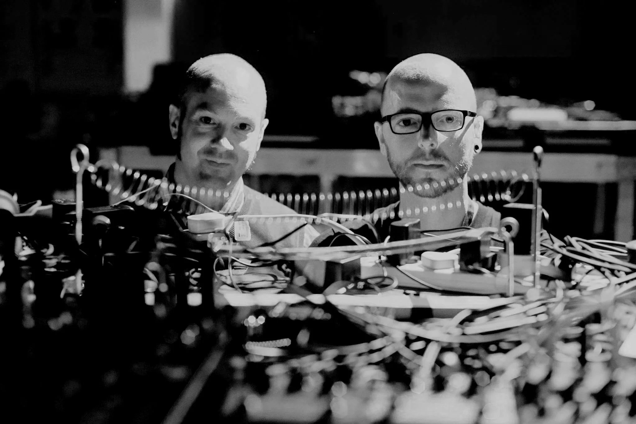 Two men with shaved heads and glasses behind a cluttered electronic project setup at night.