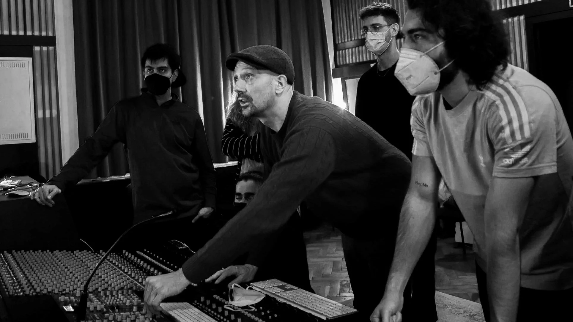 A group of people in a recording studio working with audio equipment, all wearing face masks. One person is operating a mixing console, while others observe.