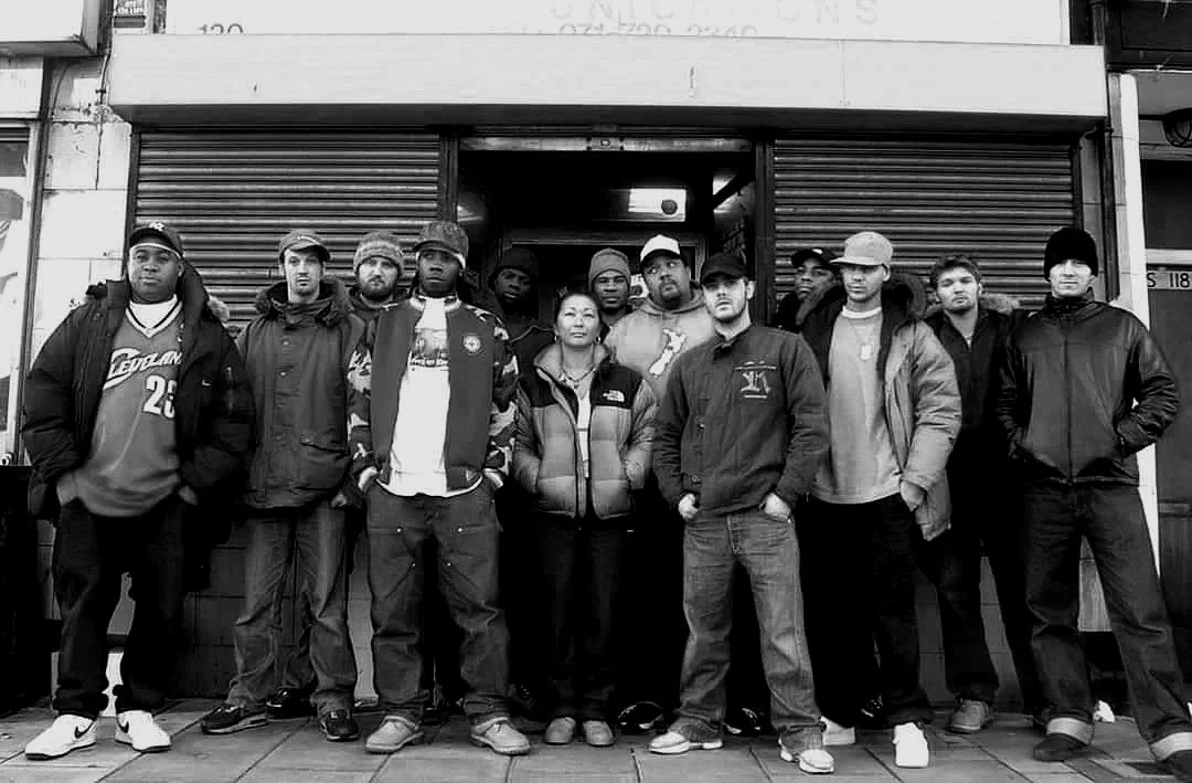Group of fifteen diverse people standing in front of a closed storefront on a city street, wearing casual clothing and jackets.