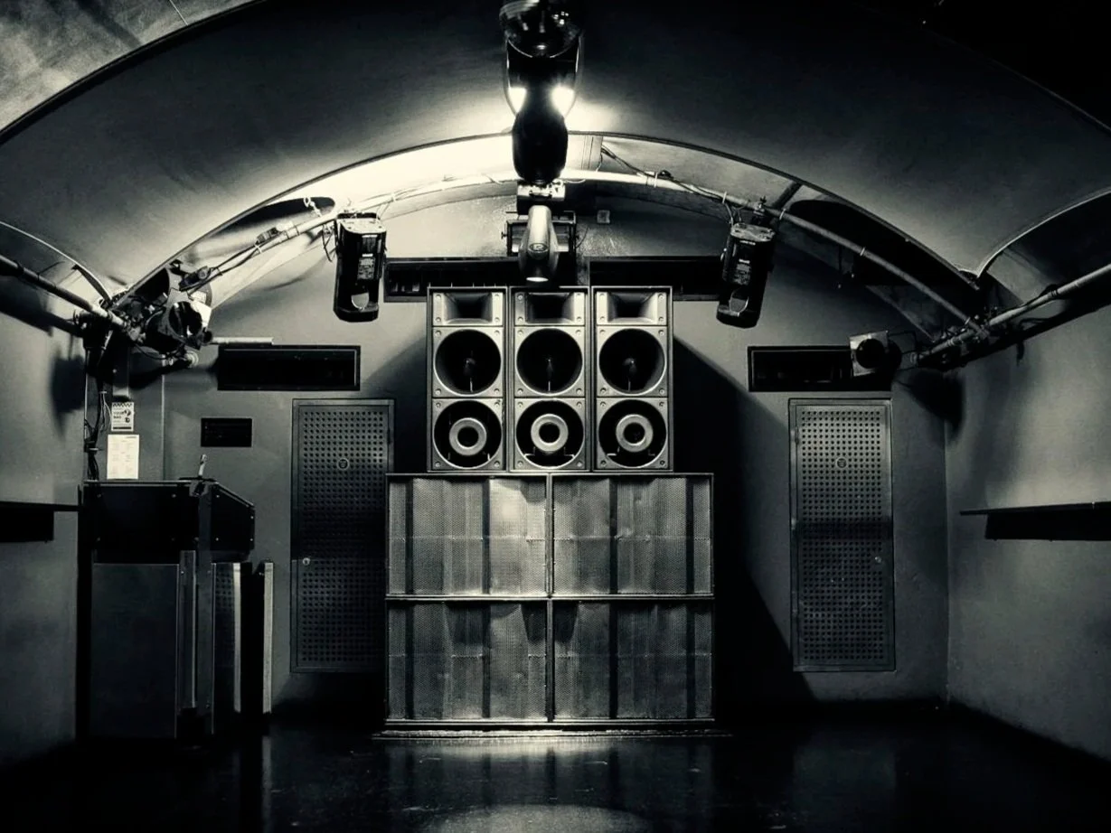 A dark room with a curved ceiling, featuring large speaker arrays mounted on the back wall and smaller speakers and equipment hanging from the ceiling.
