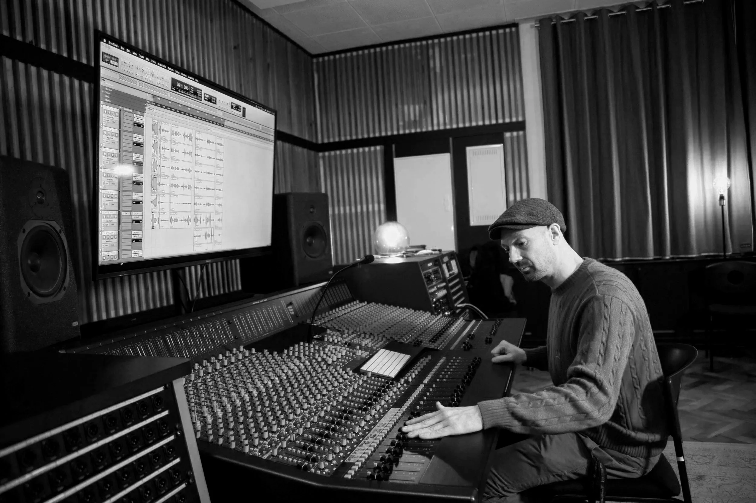 A person with a beanie and sweater working with audio equipment in a recording studio, viewed from the side with a large computer screen displaying audio tracks, speakers, and a mixing console.