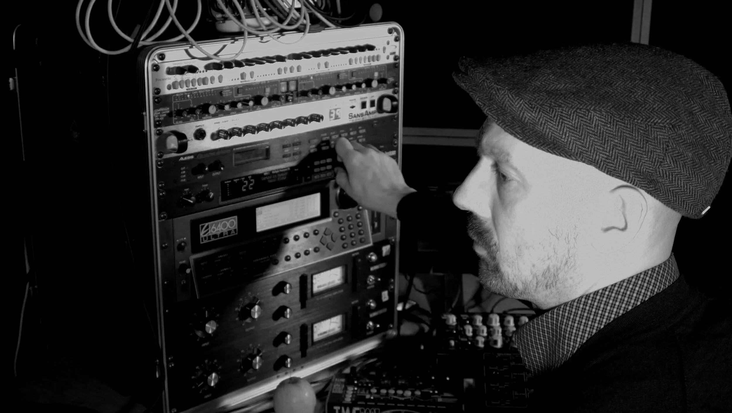 A person wearing a flat cap is adjusting audio equipment with various dials and controls, in a monochrome setting.