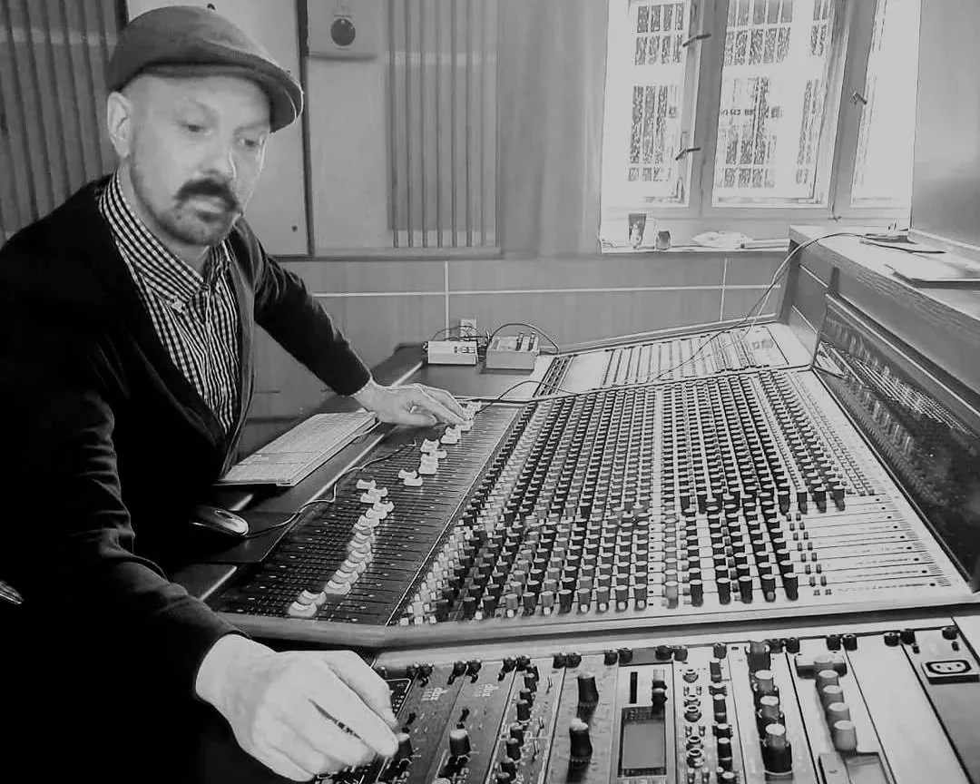 Black and white photo of a man with a mustache, beard, and hat operating a large sound mixing console.