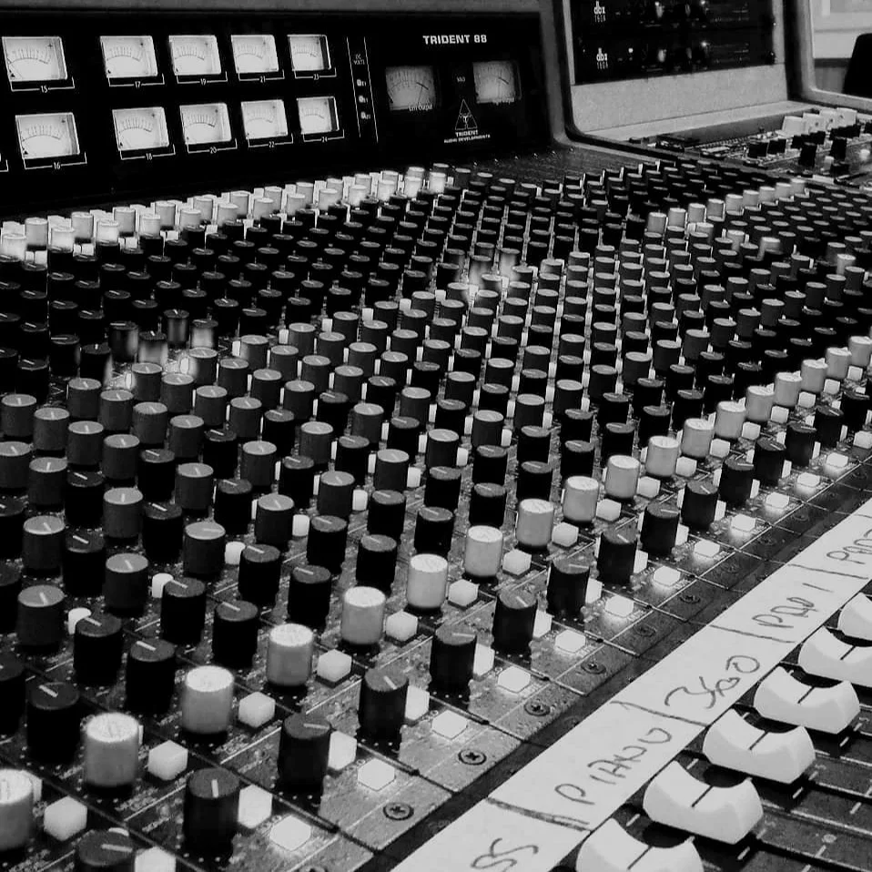 Black and white photo of a professional audio mixing console with numerous knobs and sliders, and a handwritten note partially visible at the bottom.