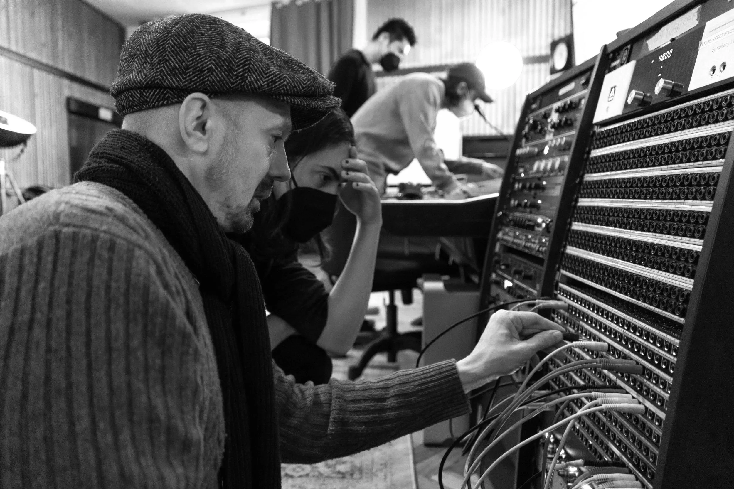 Group of people working on audio equipment, focusing on a man adjusting wires connected to a large audio mixer in a room with wood-paneled walls.