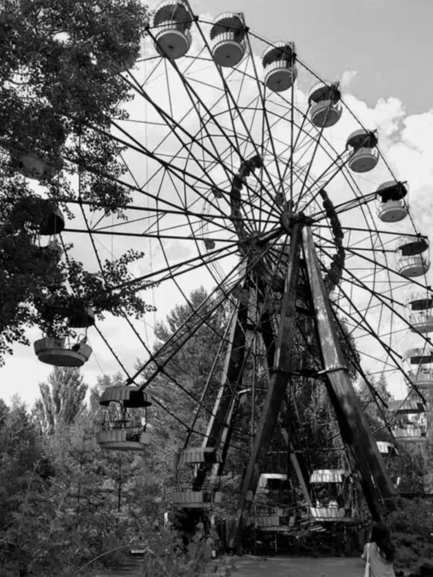 Black and white photo of a large Ferris wheel in a park with trees around it, some rides appear to be old and unused.