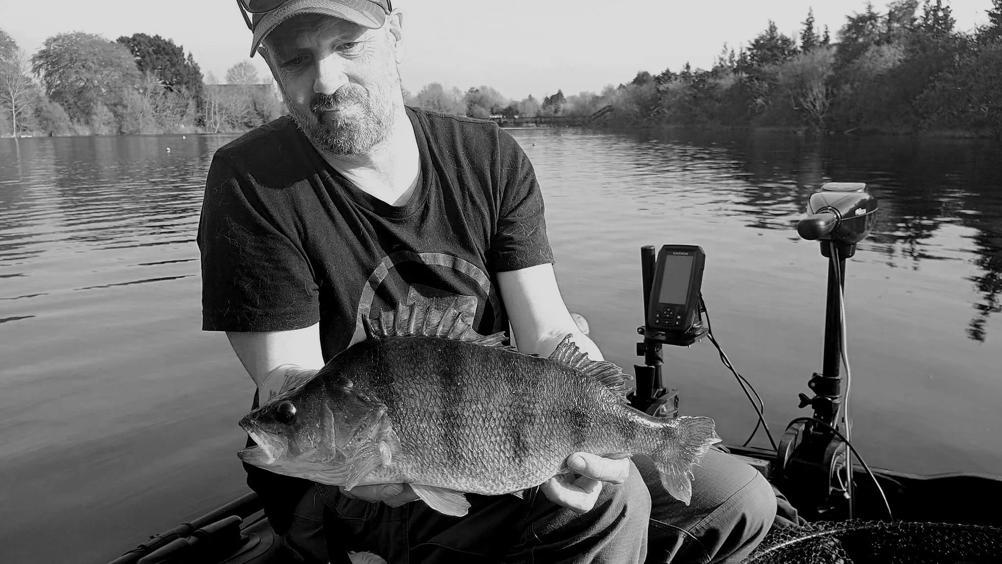 A man with a beard and cap holding a large fish on a boat, with a fishing rod and fishfinder nearby, on a calm river with trees in the background, in black and white.