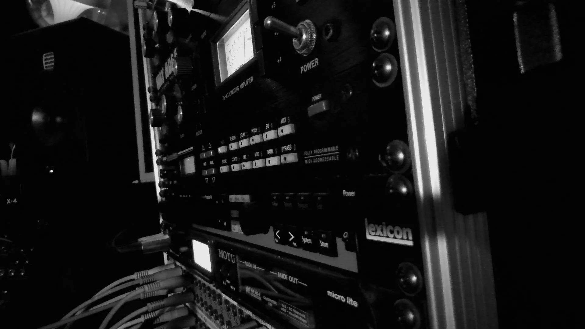 Black and white image of audio equipment, including a DJ mixer or synthesizer with knobs and buttons, connected by numerous cables.