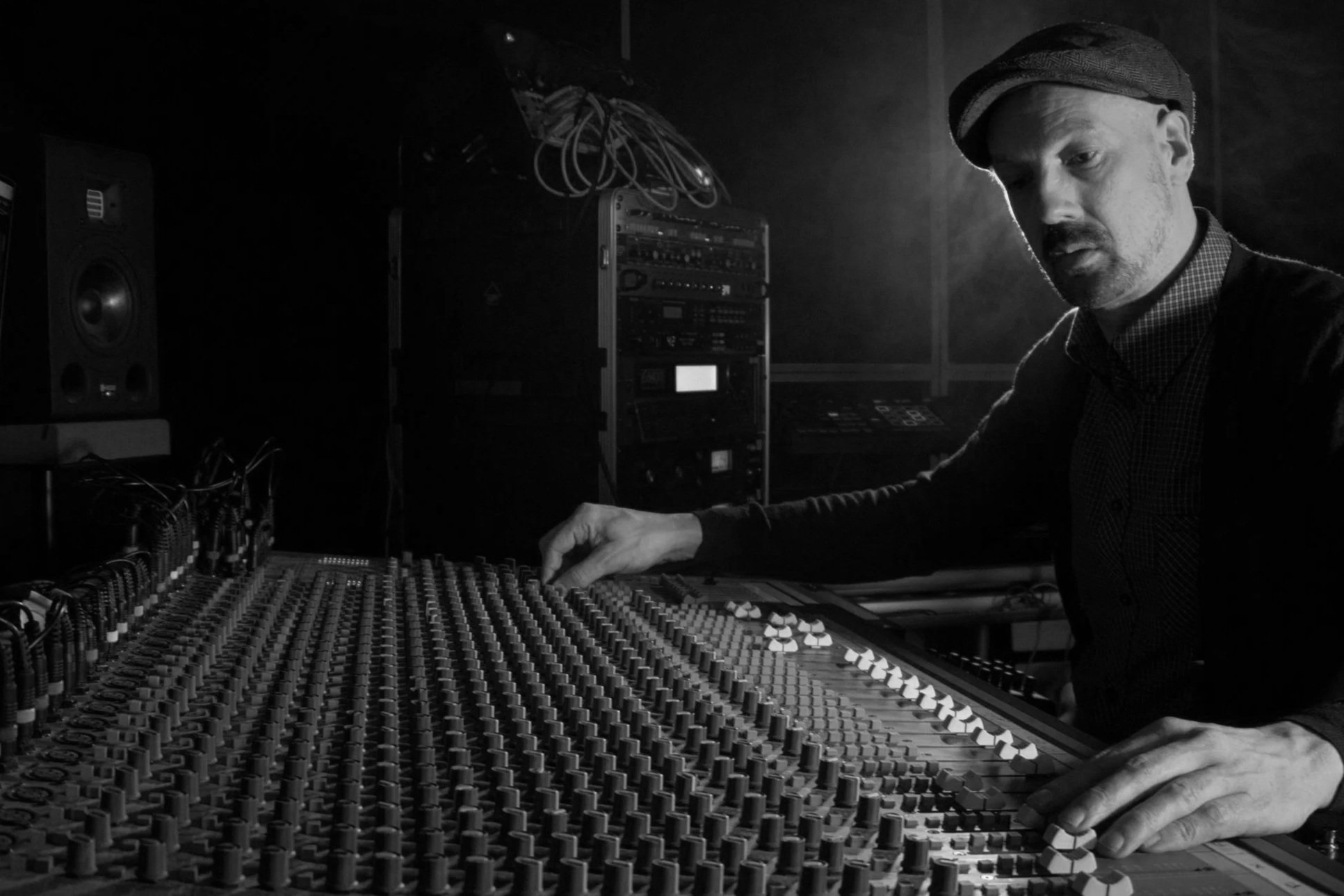 Black and white photo of a man wearing a cap working with audio equipment in a studio, adjusting controls on a large audio mixing console.