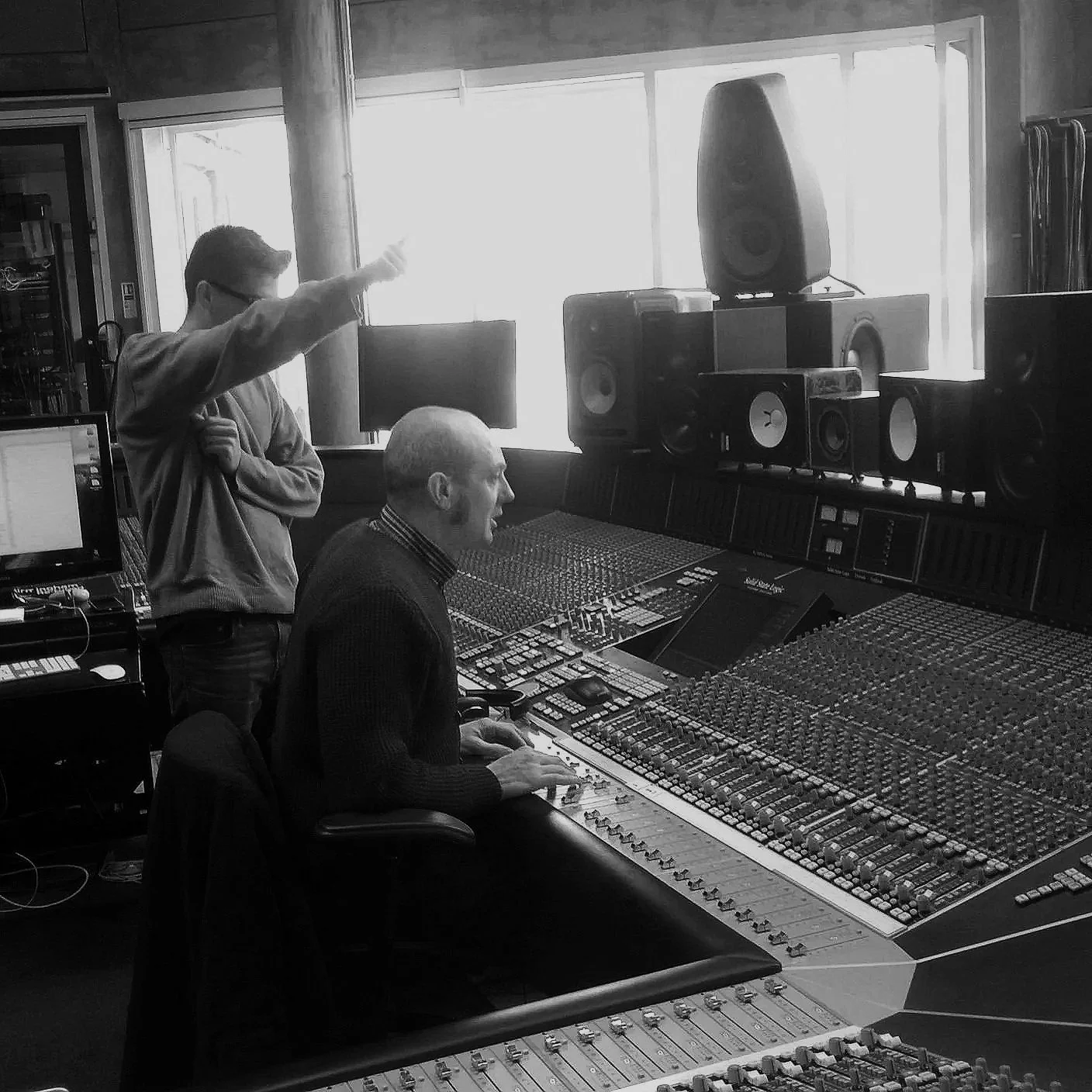 Two men in a recording studio; one is sitting at a mixing console, and the other is standing and pointing towards the window behind a large speaker.