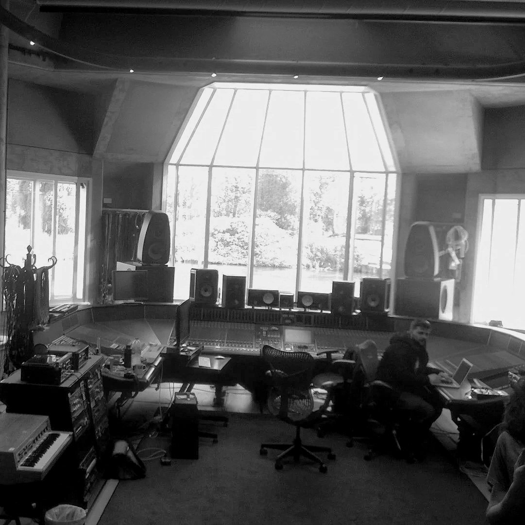 A modern recording studio with large floor to ceiling windows overlooking a landscape with trees. The studio has audio equipment, mixing consoles, speakers, and computers. A person is working at a computer desk, and another is seated nearby.