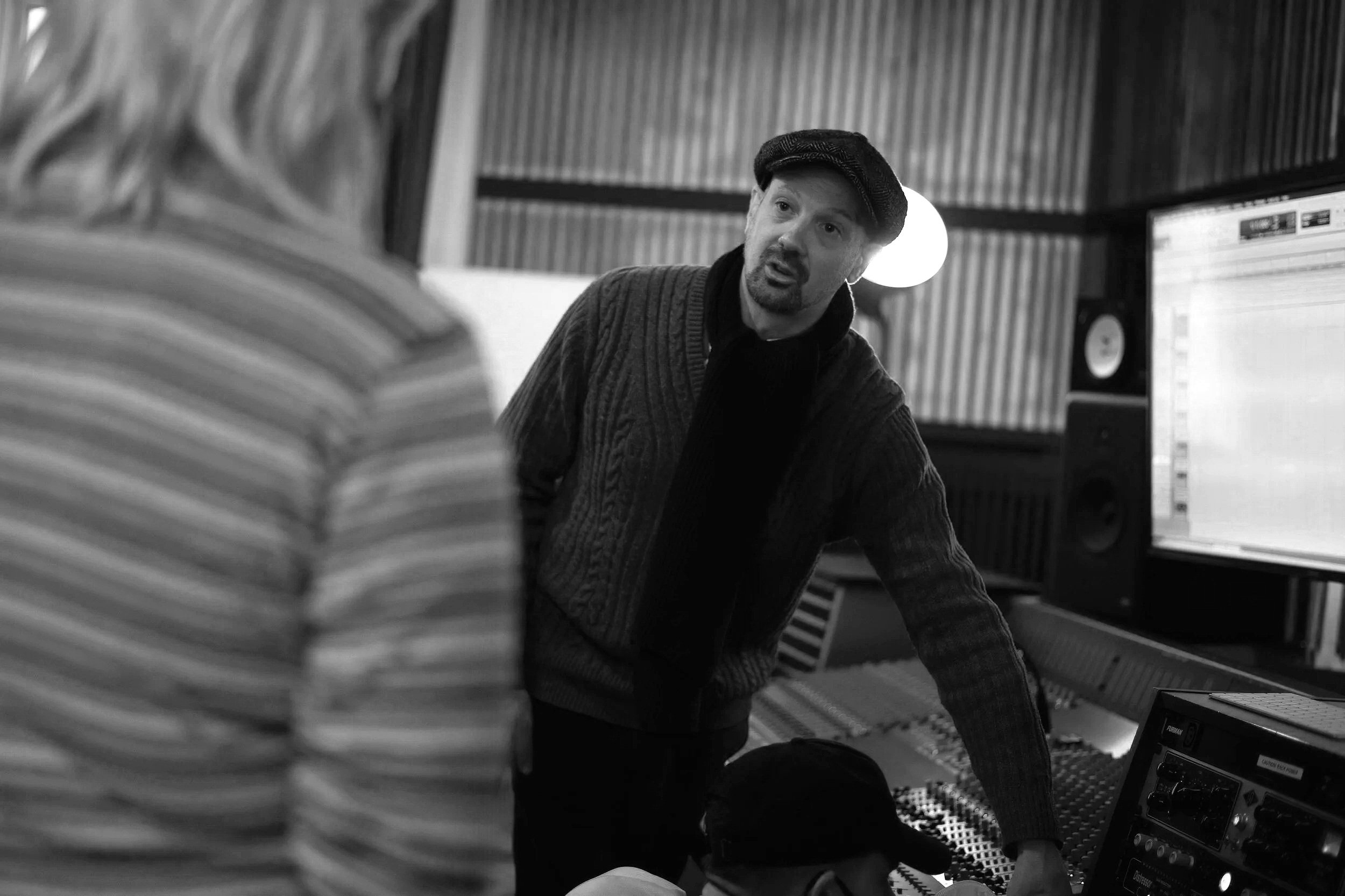 A man with a beard and wearing a flat cap is leaning over a mixing console or studio equipment in a recording or music studio, engaged in a discussion or explanation.