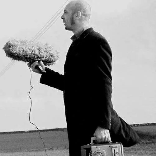 A man in a suit holding a large object resembling a drone with a fluffy exterior, along with a briefcase, standing against a plain wall.