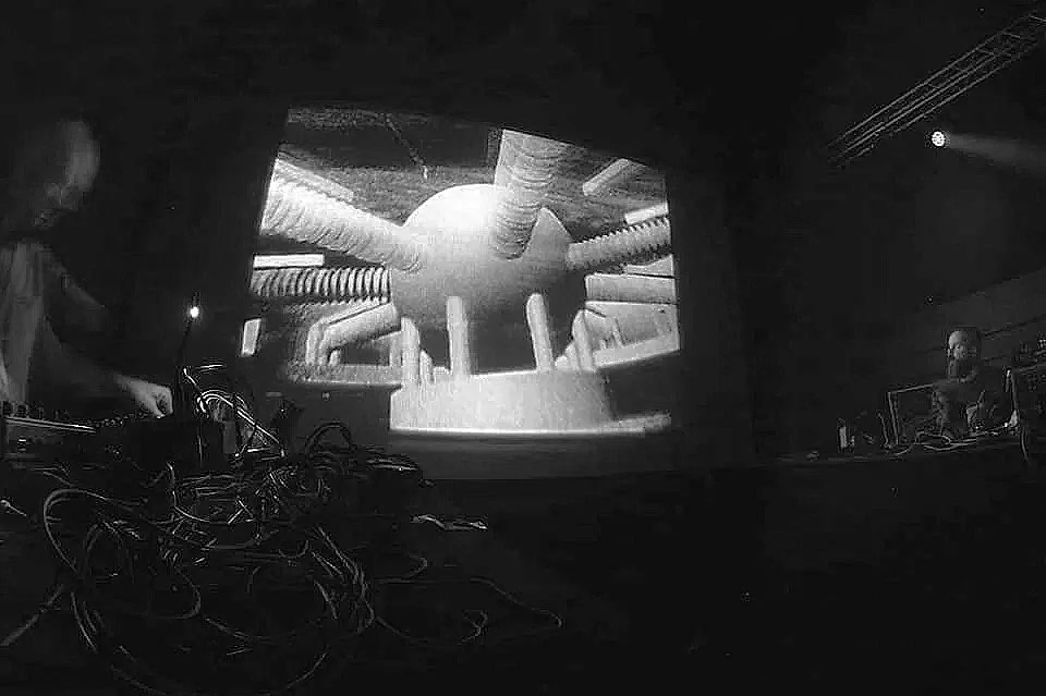Black and white image of a large projector screen displaying a computer-generated image of a futuristic machine with tubes and pipes. There are two individuals on either side of the screen operating equipment, with various wires tangled in the foreground.