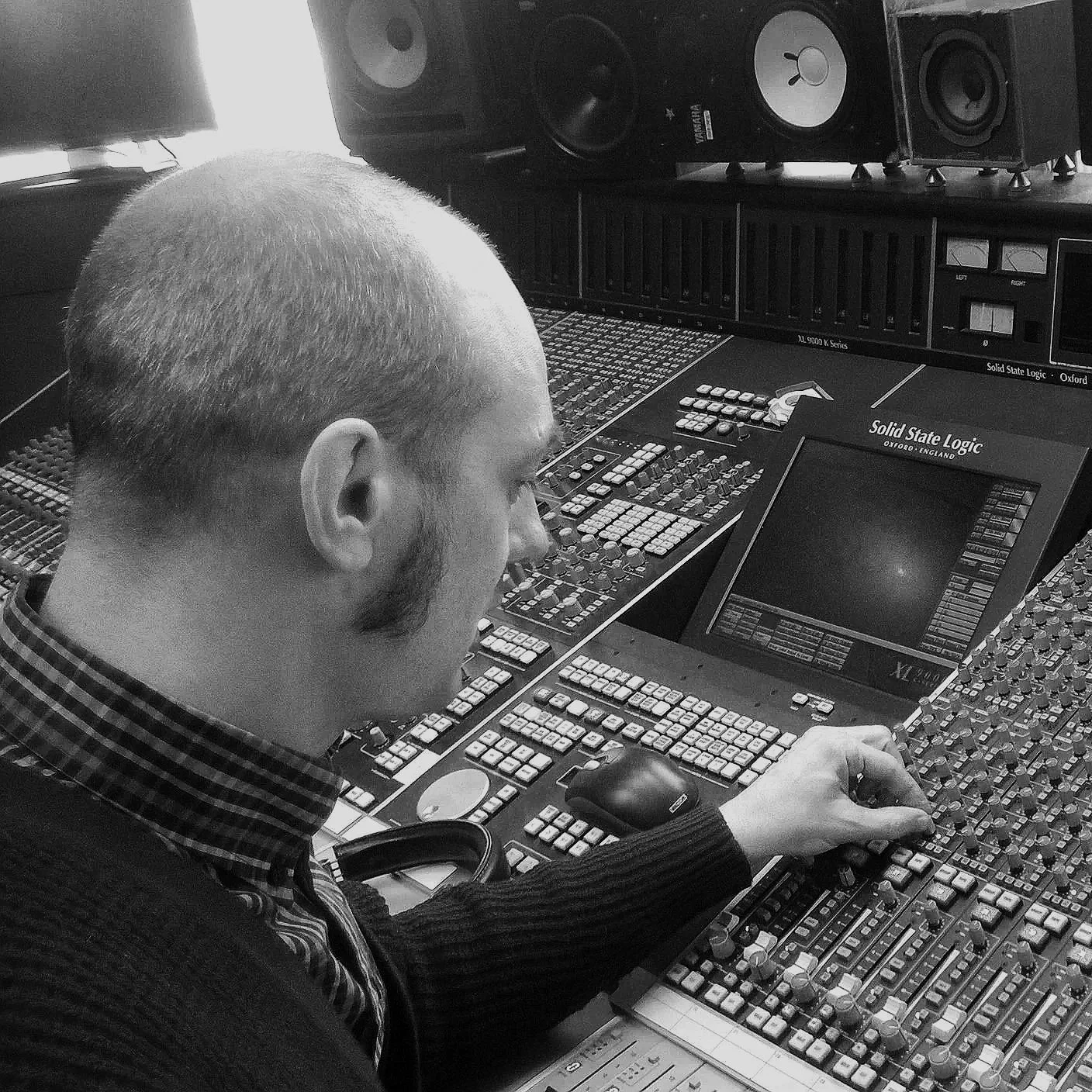 A man with short hair and a beard, wearing a plaid shirt and a dark sweater, working at a professional audio mixing console with multiple buttons, sliders, and a screen, surrounded by studio speakers.