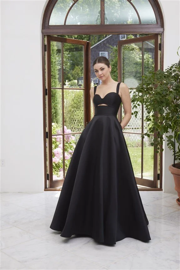 A woman in a black formal gown standing inside near open glass doors, with a garden outside.