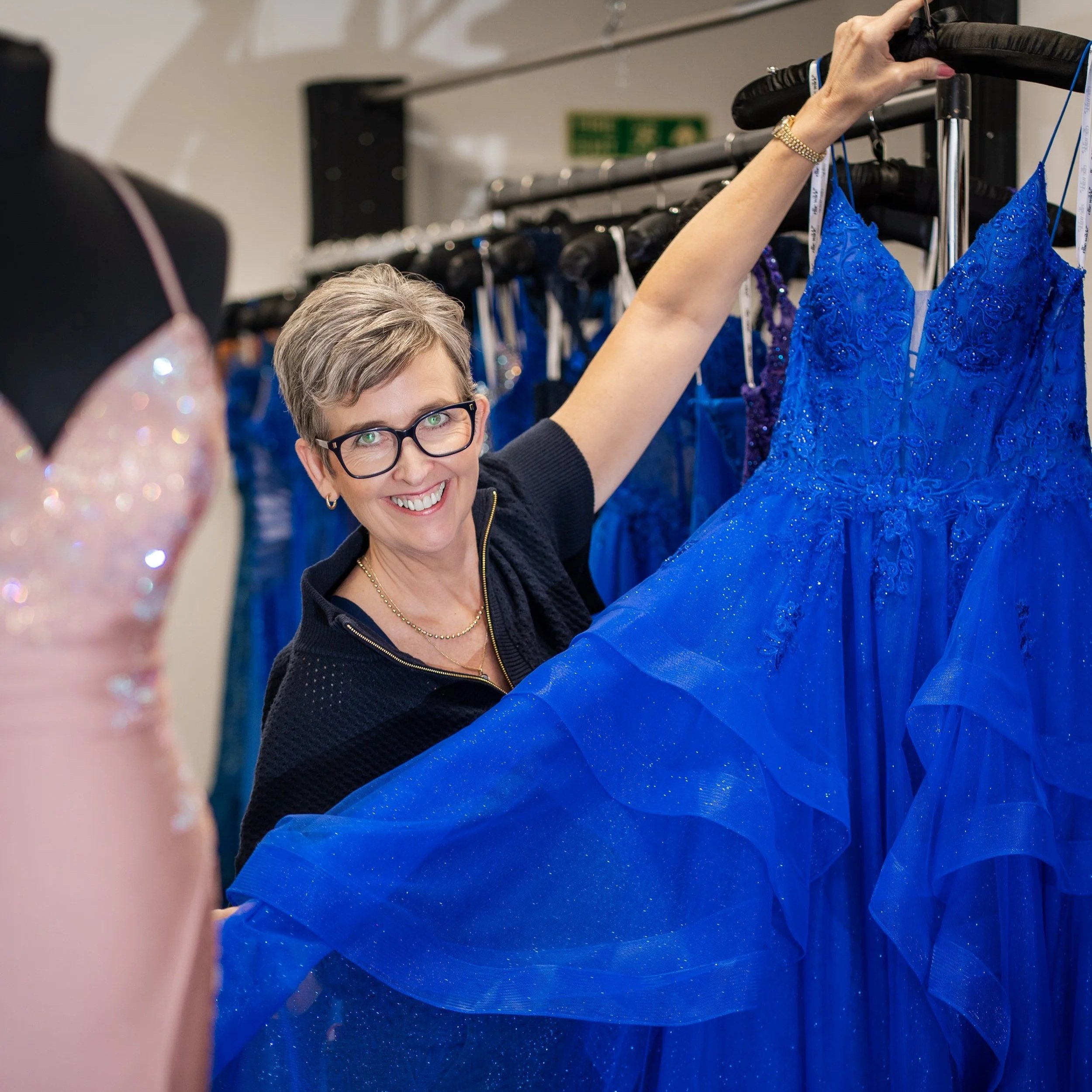 A woman with short hair and glasses is smiling while holding up a blue, beaded dress on a hanger among other dresses on a rack.
