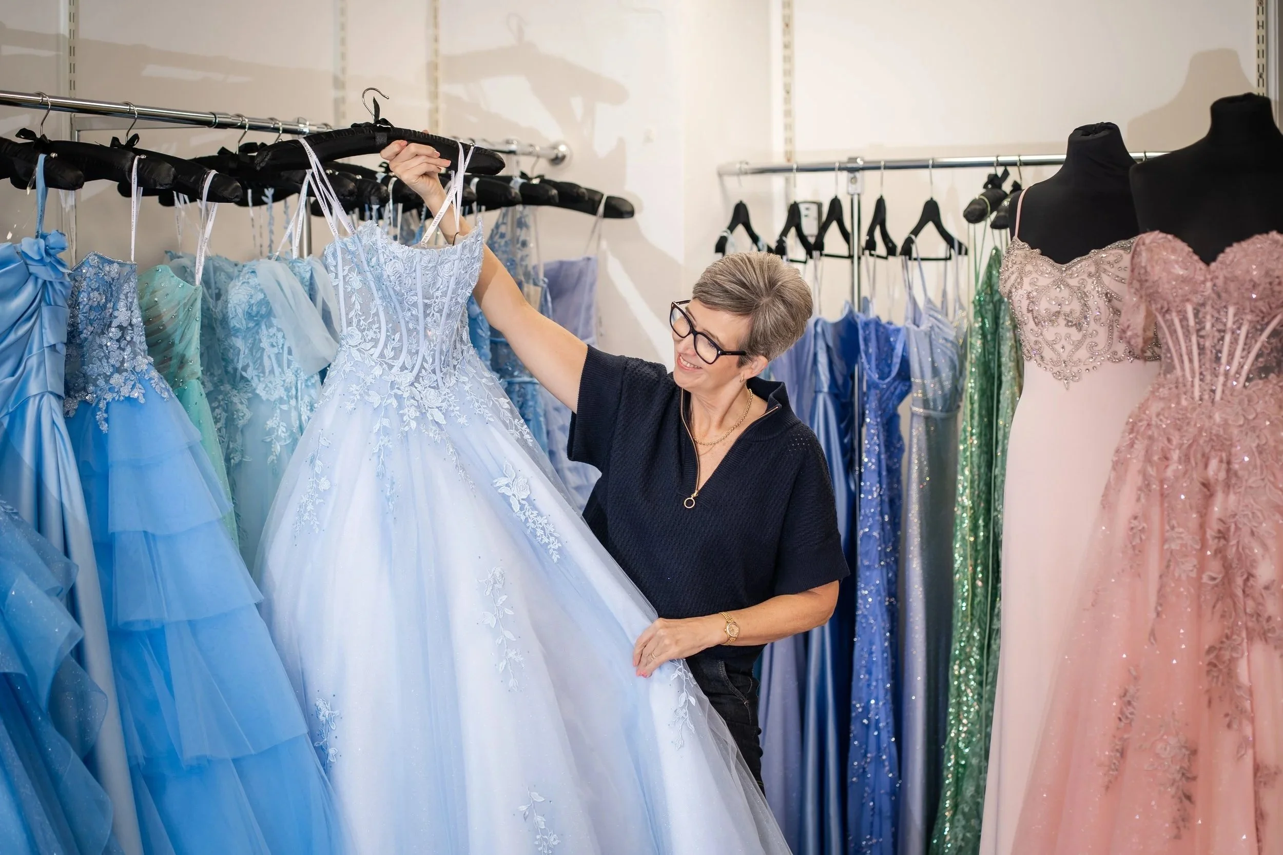 A woman shopping for dresses in a formal dress boutique, holding a light blue gown with lace details, surrounded by other colorful formal dresses and mannequins.