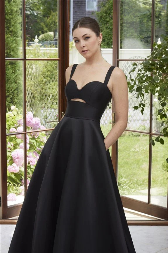 Young woman in a black evening gown standing indoors in front of open glass doors with a garden view.