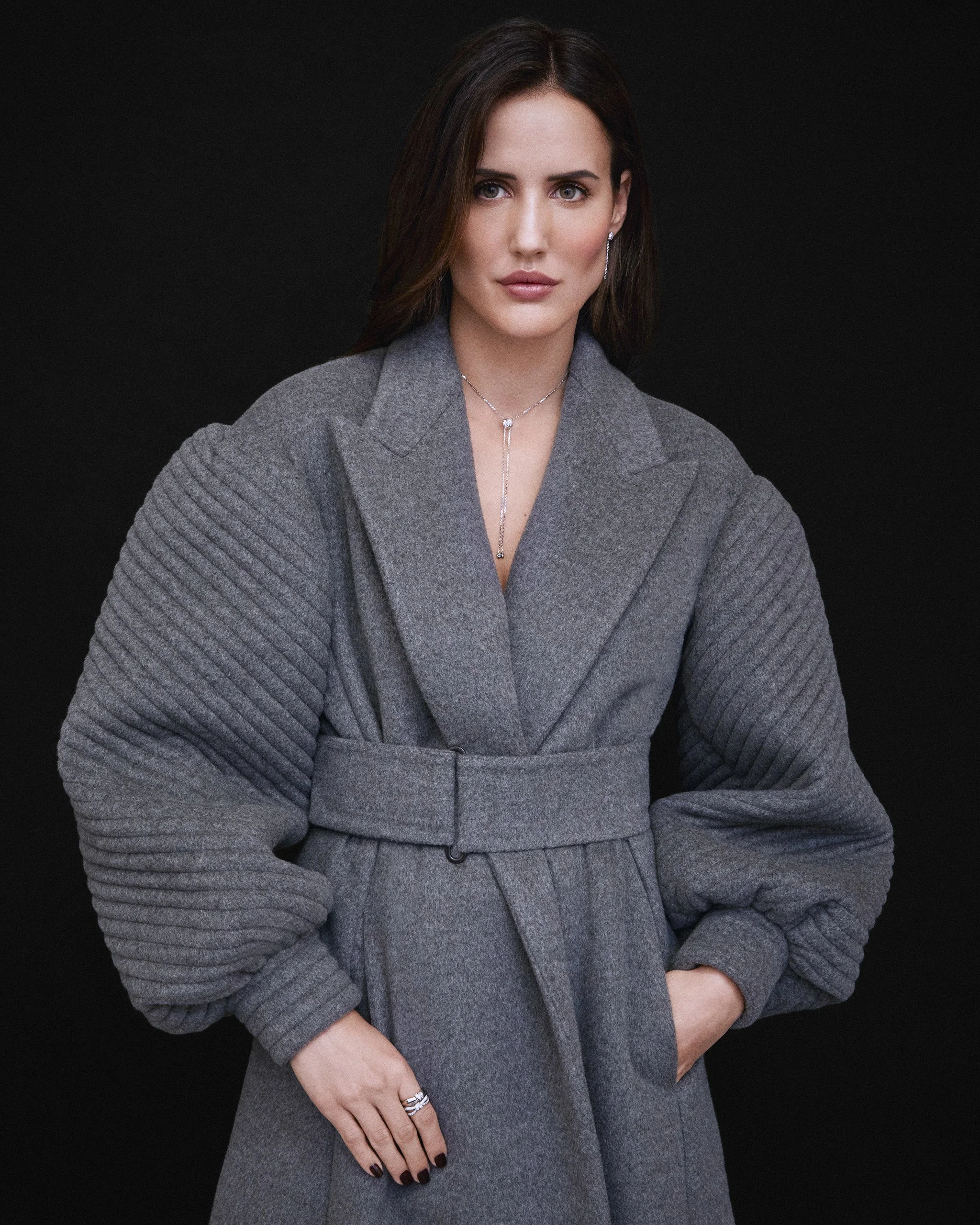 Mujer con cabello castaño oscuro, vestido gris, fondo negro, mirando a la cámara.