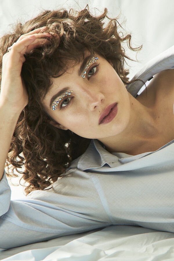 Mujer con cabello rizado y maquillaje con piedras brillantes cerca de los ojos, descansando sobre una superficie blanca.