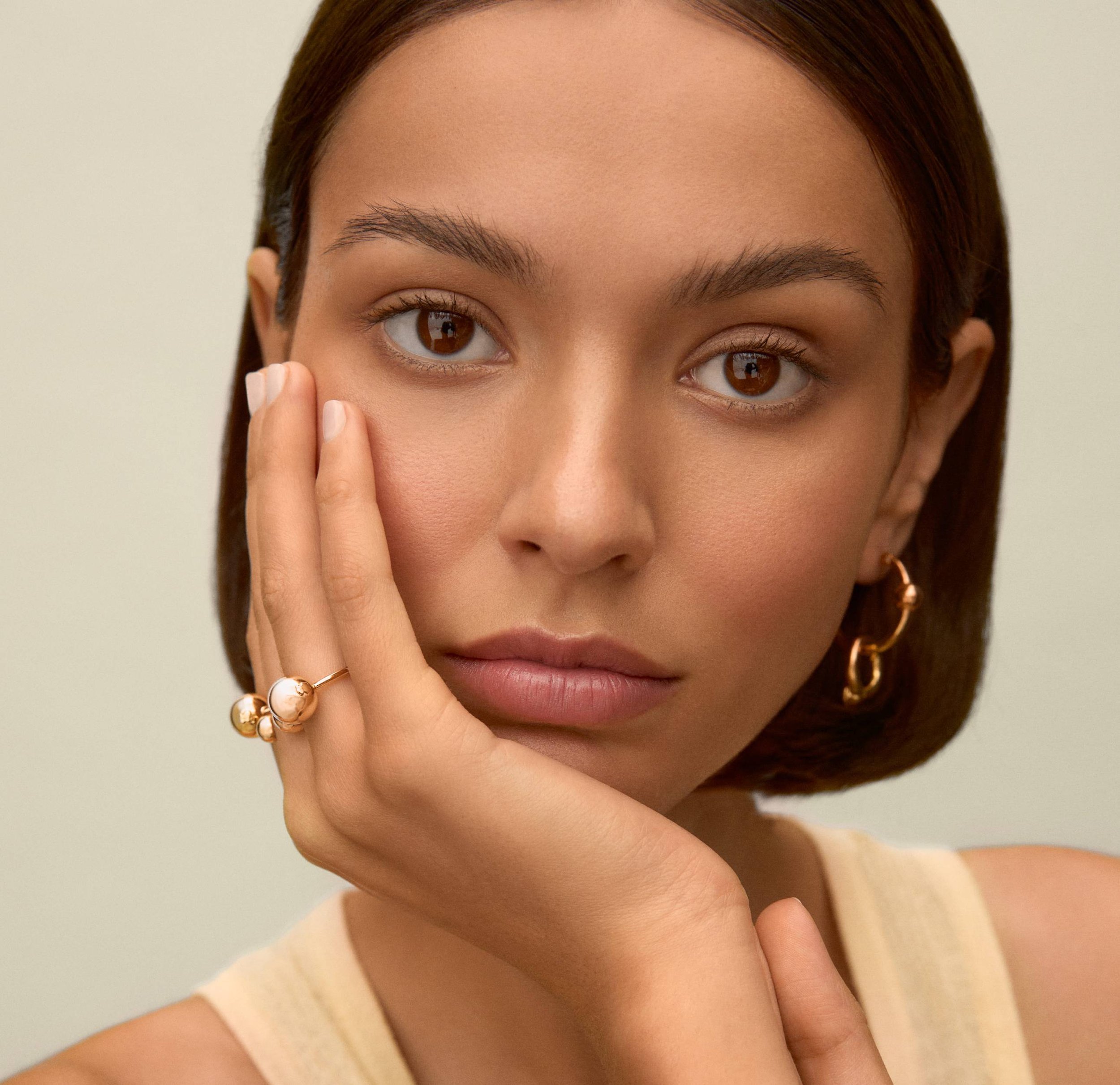 Retrato de una mujer joven con piel clara, ojos marrones, cabello castaño y suelto, usando aretes dorados y anillos en sus dedos. Ella está tocando su mejilla con su mano izquierda y mirando hacia adelante.
