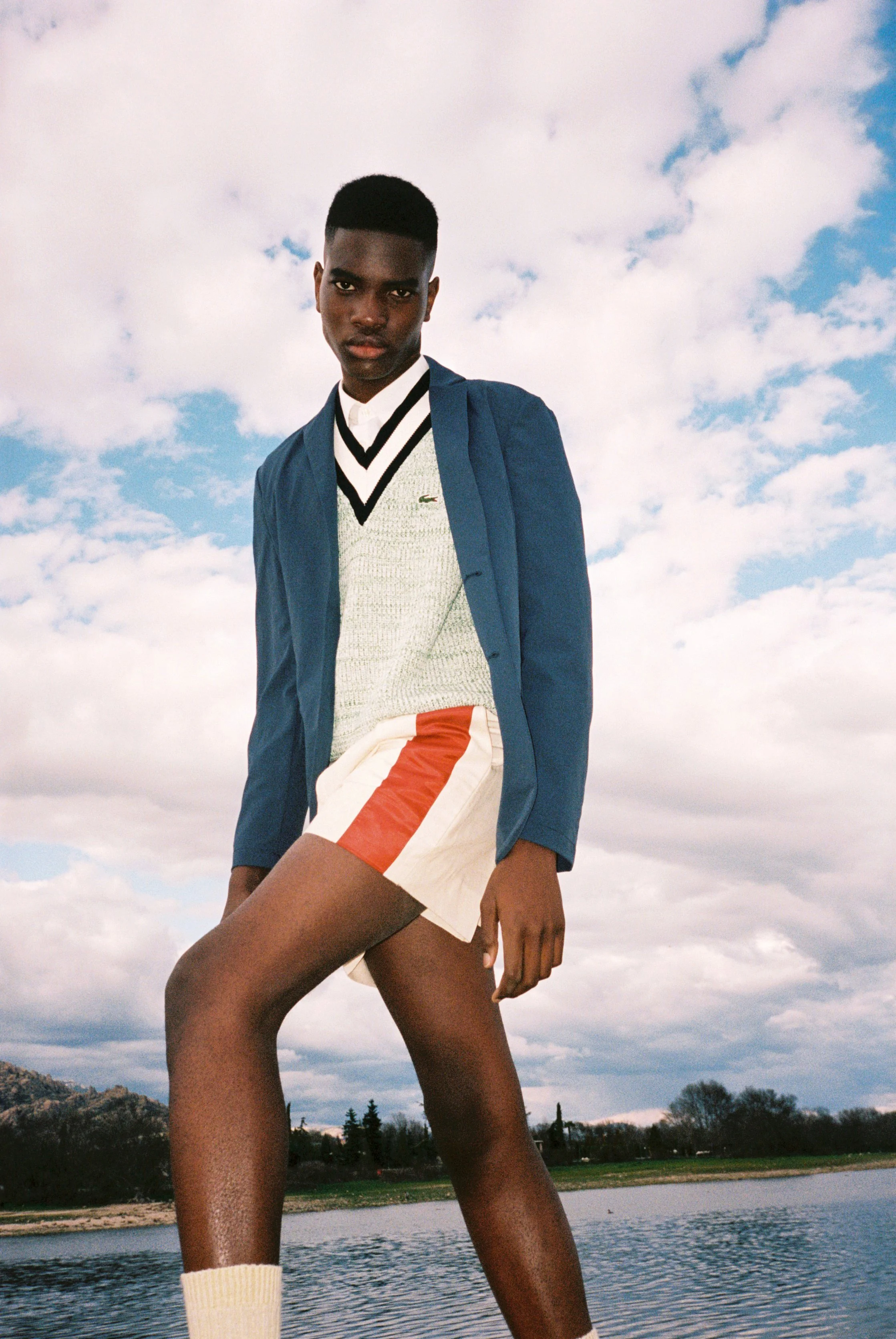 Joven de piel oscura, vistiendo una chaqueta azul, un suéter gris con detalles en blanco y negro, una camisa blanca, pantalones cortos de color beige con detalles en rojo y blanco, en un entorno al aire libre con un cielo nublado y un cuerpo de agua en el fondo.
