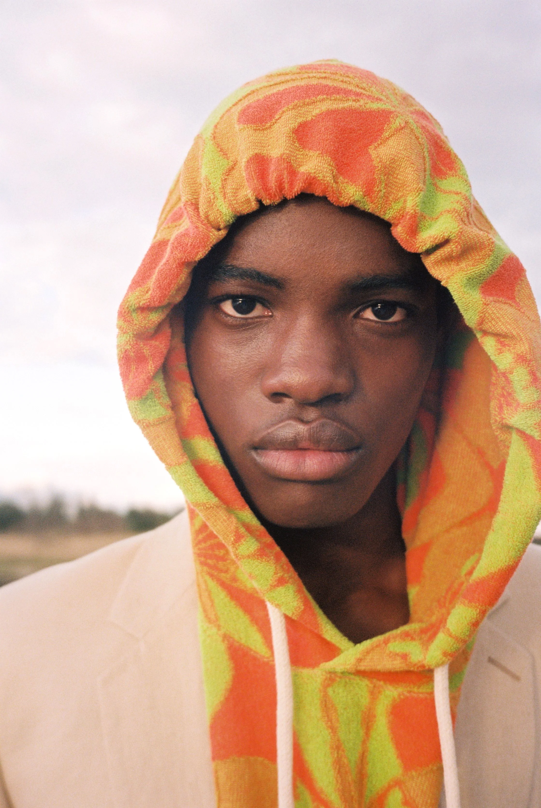 Joven con piel oscura usando sudadera con capucha de colores naranja, amarillo y verde, mirando directamente a la cámara, contra un cielo nublado de fondo.
