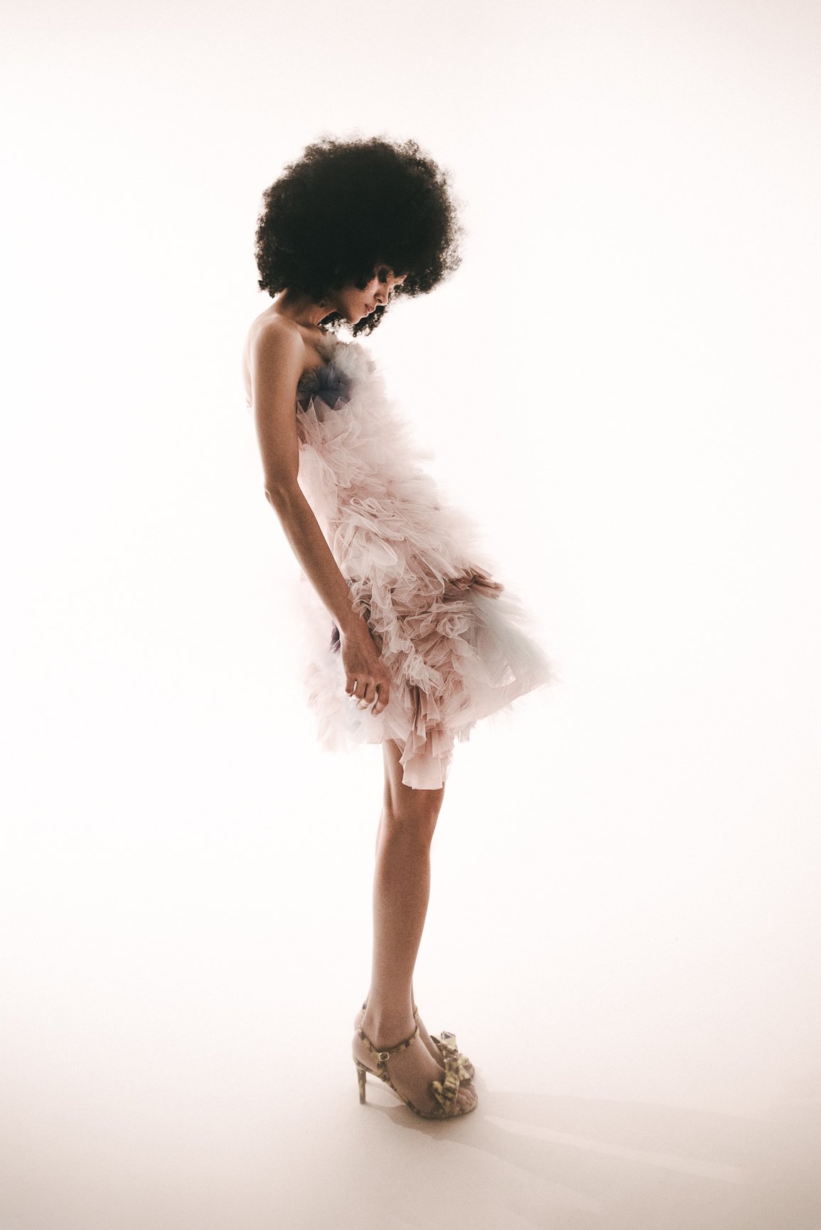 Una mujer con un vestido rosa de tul y tacones dorados posa de perfil con la cabeza agachada.