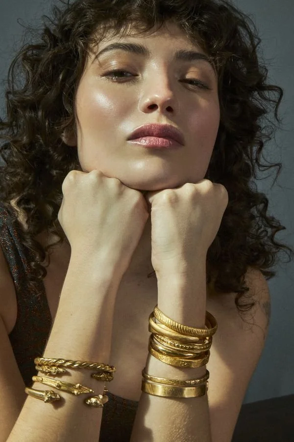 Mujer con cabello rizado, usa varias pulseras doradas y mira hacia la cámara con expresión relajada.