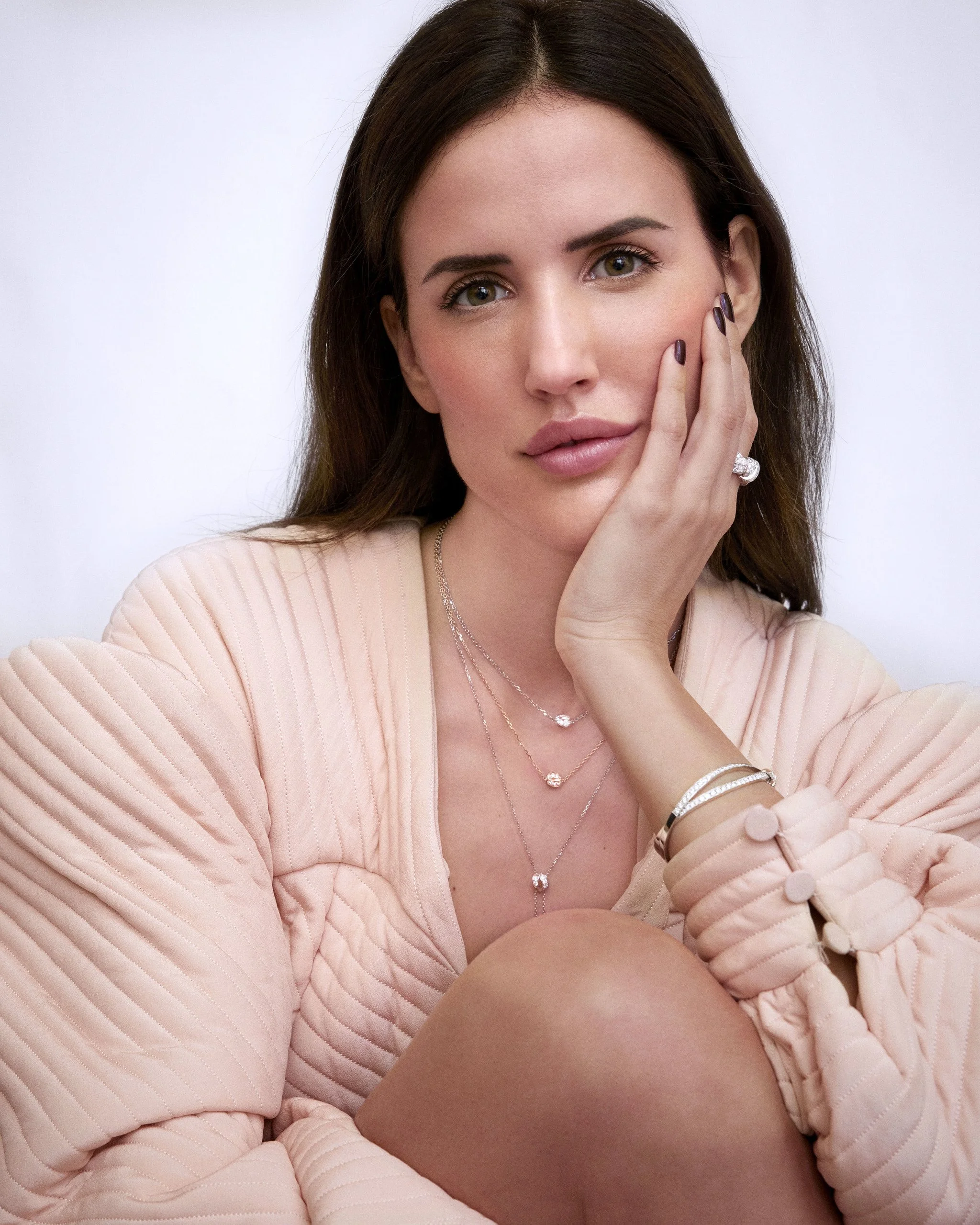 Retrato de una mujer joven con cabello castaño, piel clara y ojos verdes, luciendo un conjunto de joyería y un suéter color rosa claro, con expresión pensativa y la mano en la mejilla.
