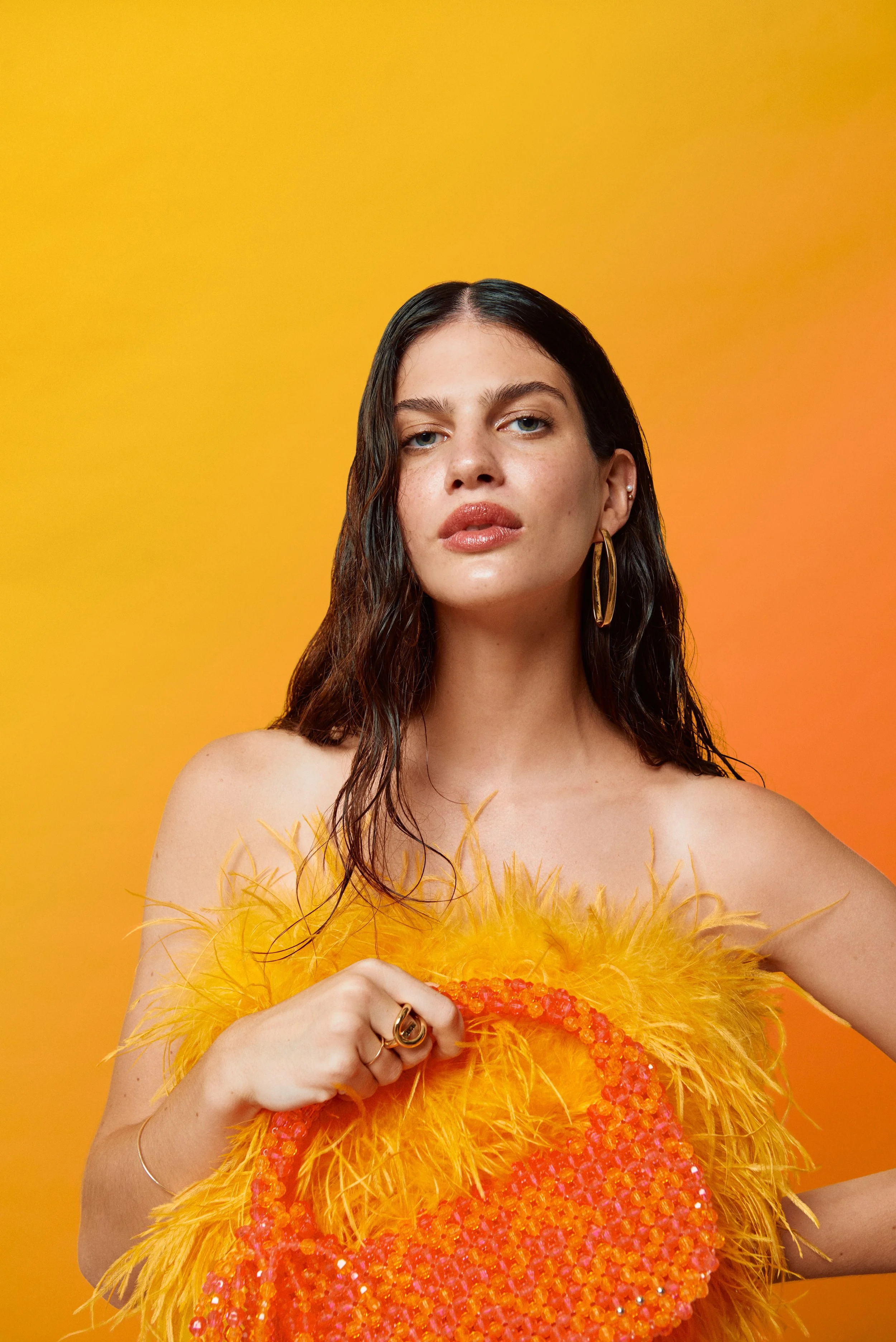 Mujer con vestido de plumas amarillas y bolso de cuentas naranjas, fondo amarillo y naranja.