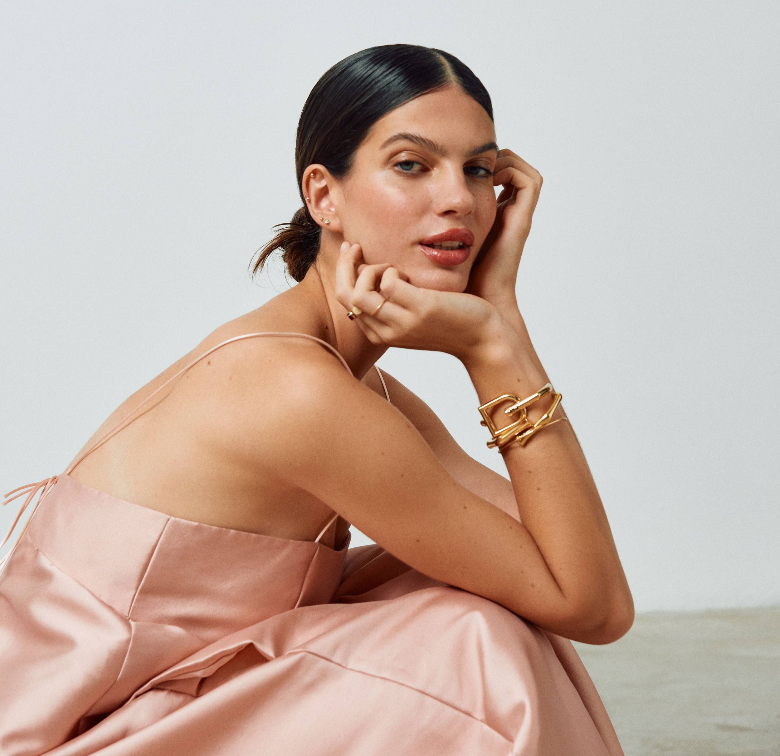 Retrato de una mujer con piel clara, vestida con un vestido de color rosa pálido, con cabello oscuro recogido, usando aretes dorados, pulseras y anillo, posando con la cabeza apoyada en una mano y mirando hacia la cámara.