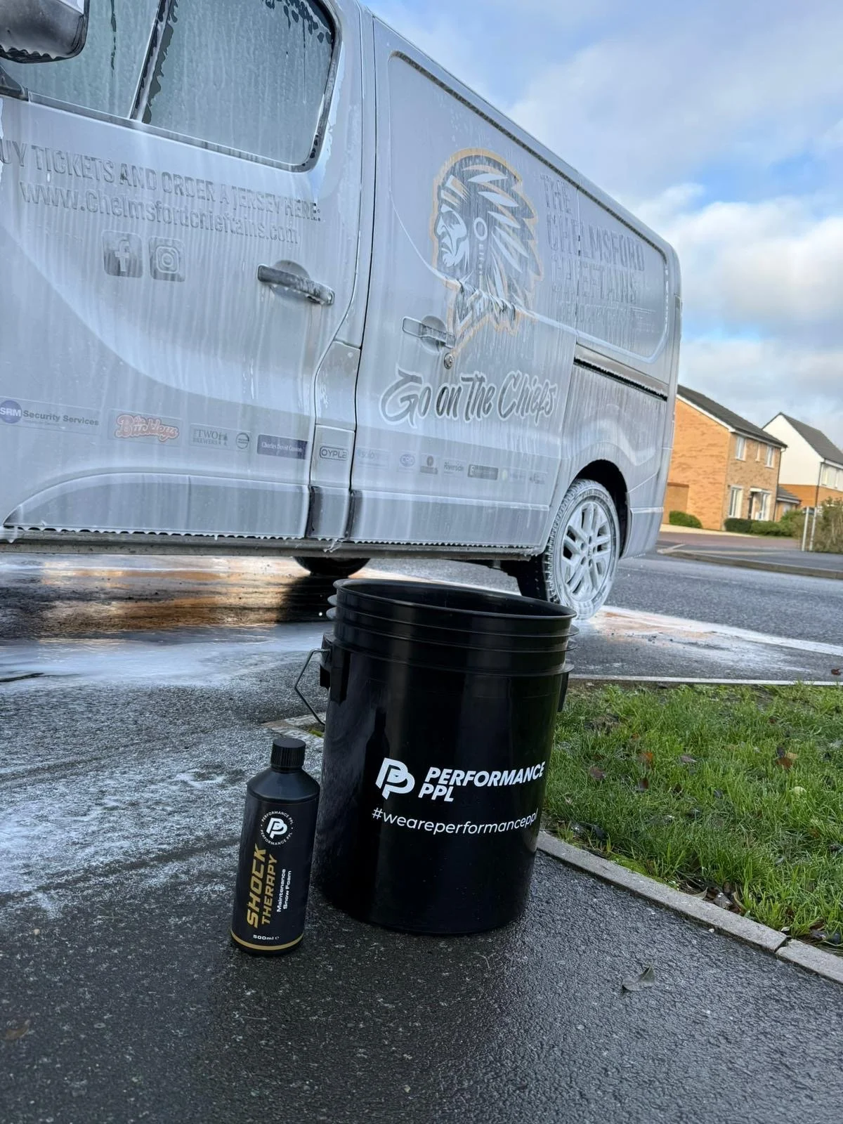 A black bucket labeled 'Performance PPL' with the hashtag '#weareperformanceppl' sits on wet pavement next to a bottle labeled 'Shock Therapy,' with a lock attached to the bucket handle. In the background, a gray van with advertising and a lion logo is parked in a residential neighborhood under a partly cloudy sky.