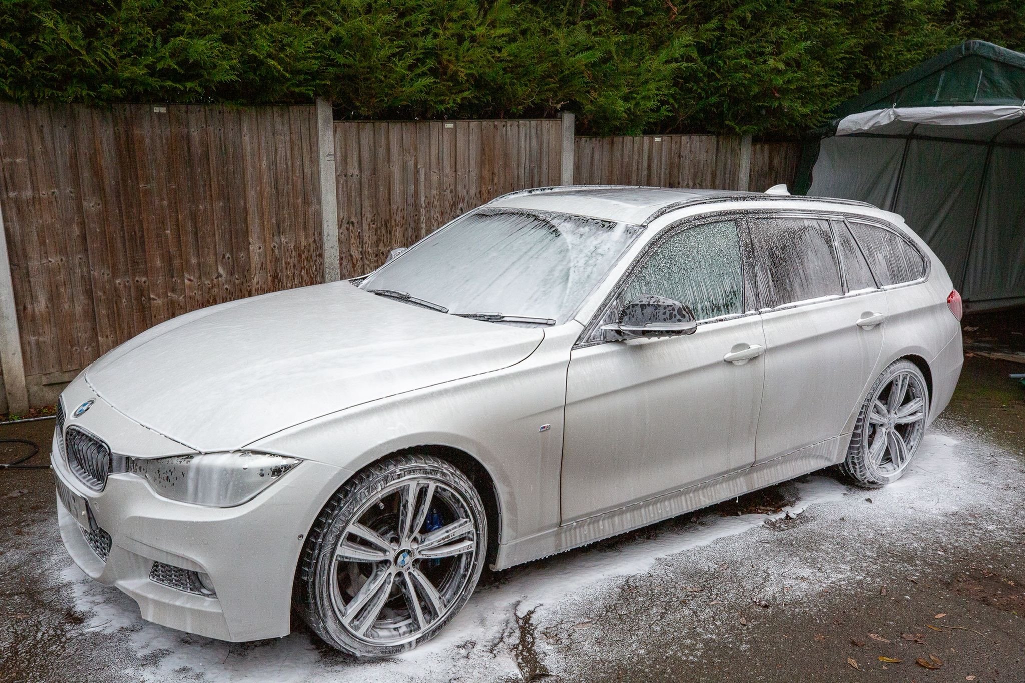 A white BMW station wagon car being washed with soap suds and foam, parked on a driveway with a wooden fence and a green shed in the background.