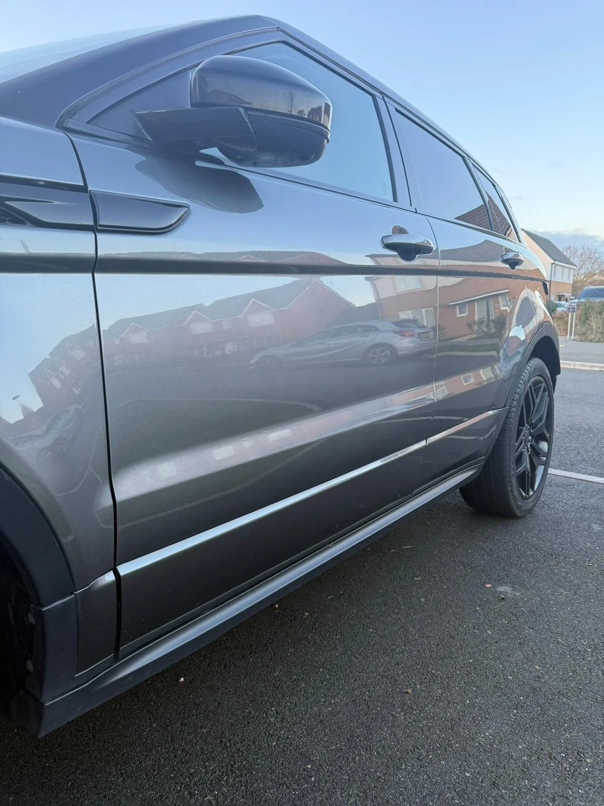 Close-up of a dark gray SUV parked on the street, showing the driver's side and reflecting nearby houses and sky on its surface.