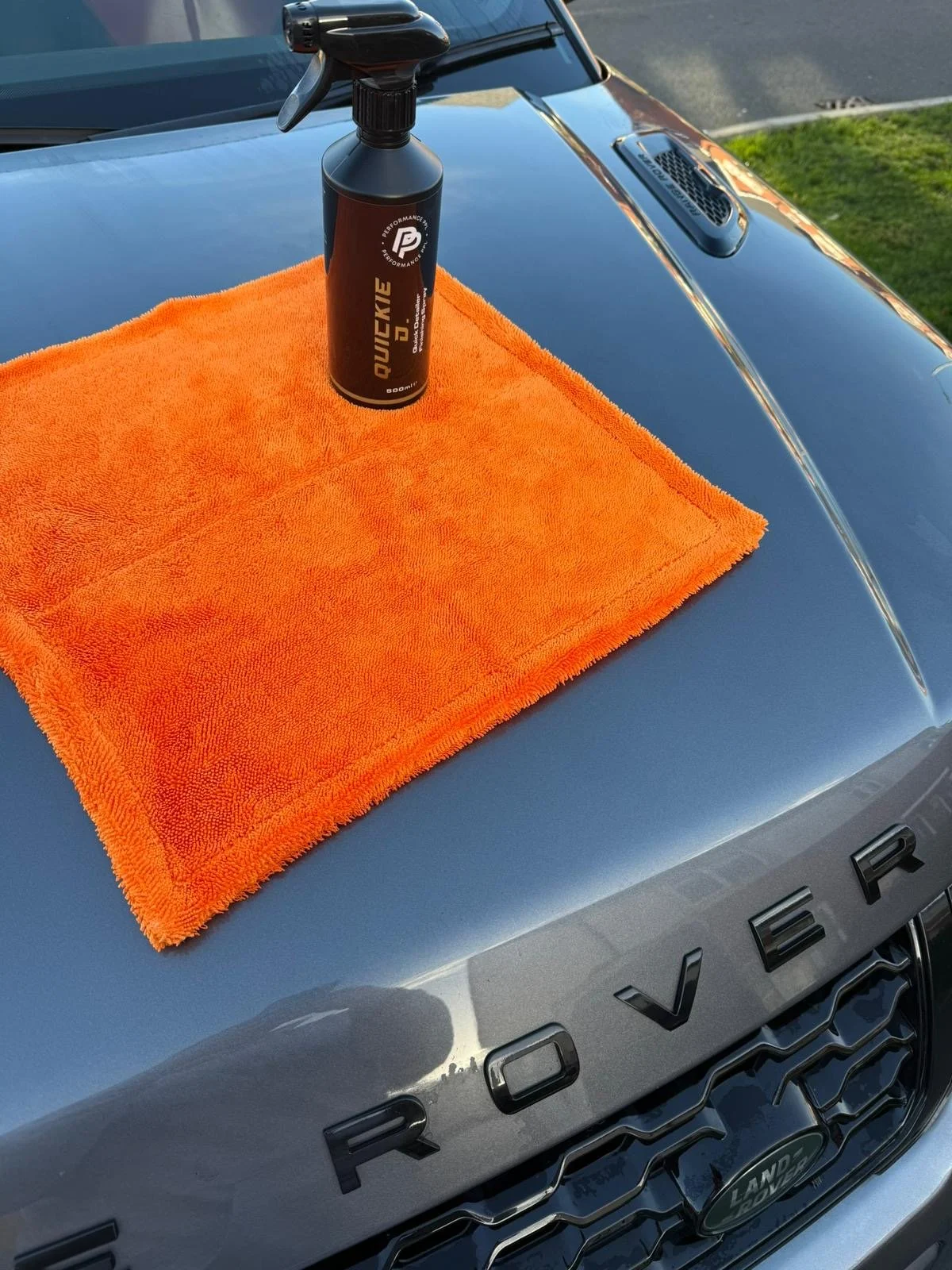 A close-up of a car hood with a black bottle of quick detail spray and an orange microfiber towel placed on it. The car is a Land Rover, and the background shows a grassy area and a street.