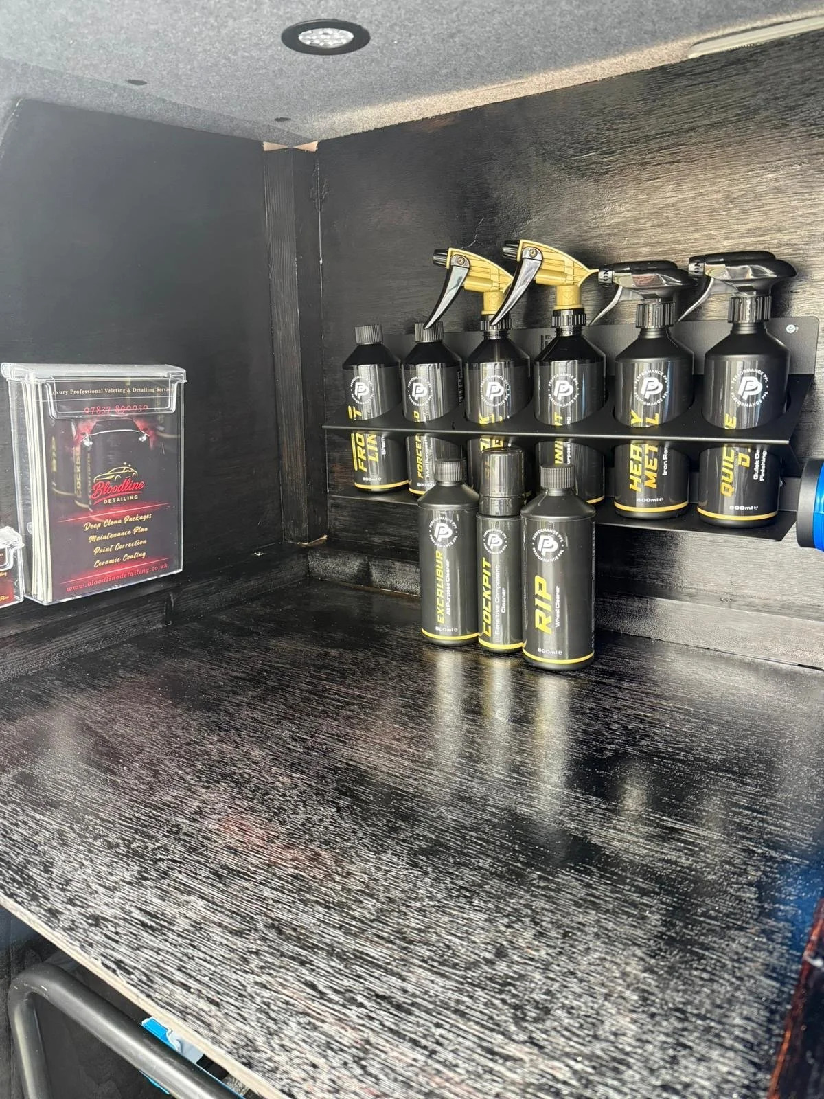 A black shelf with several black spray bottles labeled 'EXPLOSUR,' 'LOCKUP,' and 'RIP,' along with yellow spray nozzles. To the left, there is a clear plastic container with a red and black Bloodline logo and some text, mounted on the black surface.