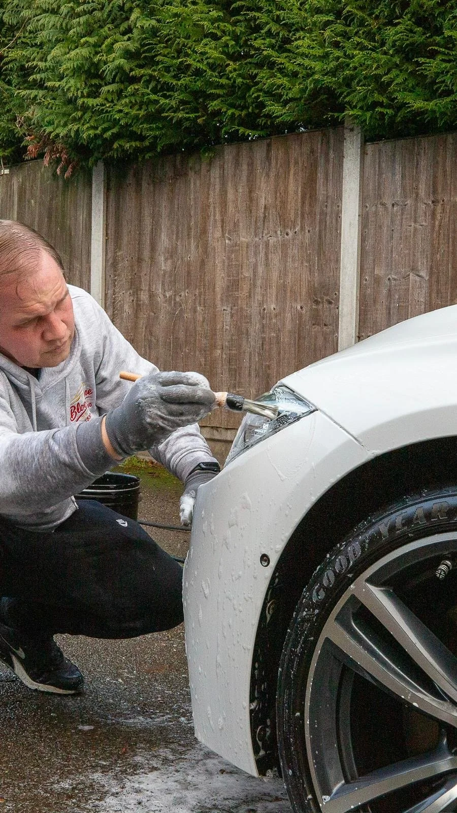 A man washing a white sports car with a brush, with soap suds on the car's front fender and tire.