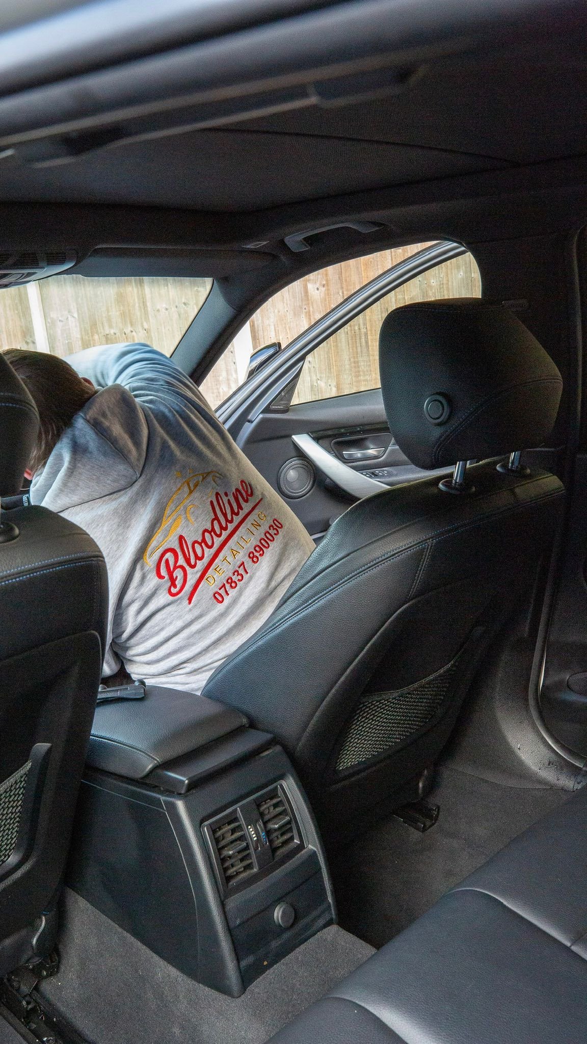 Inside the back seat of a car, showing a person leaning forward wearing a gray hoodie with red and yellow text that reads 'Bloodline Detailing' and a phone number. Car interior includes black seats, a center console with air vents, and a wooden fence visible through the window.